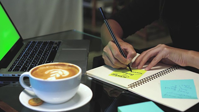 4k镜头镜头特写亚洲女商人的手工作和写在便利贴笔记本电脑与绿色屏幕笔记本电脑和喝咖啡在咖啡馆，生活方式和商业概念视频素材