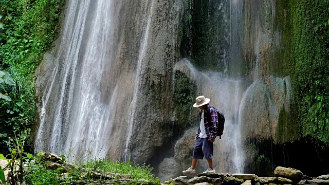 年长的亚洲人享受走在瀑布前。旅行的生活方式和成功的概念度假到野外穿着舒适的衬衫，帽子和背包。视频慢动作视频素材