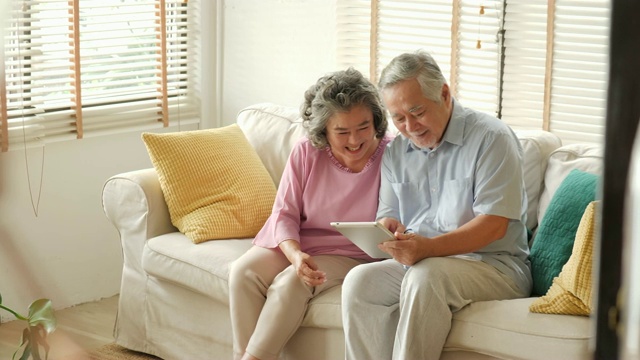 一对亚洲老年夫妇坐在家里的沙发上，用着数字平板电脑聊天、微笑。有科技观念的人。视频下载