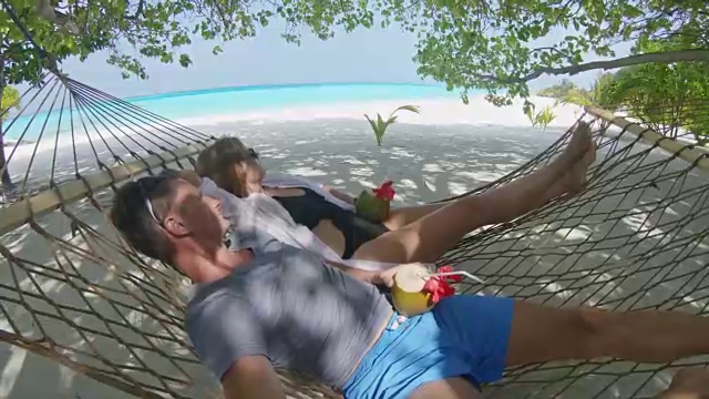 马尔代夫热带海洋海滩上，一对恬静的夫妇拿着自拍杆，喝着椰子饮料，躺在吊床上休息视频素材