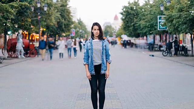 当忙碌的男人和女人四处走动的时候，一个漂亮的女孩穿着时髦的衣服看着镜头站在步行街上。视频素材