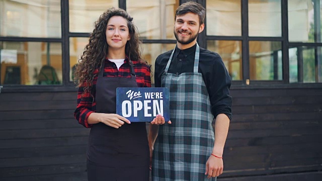 美丽的女士和她的商业伙伴英俊的男人举着开放的标志通知顾客开放他们的咖啡馆和欢迎的人的慢动作肖像。视频素材