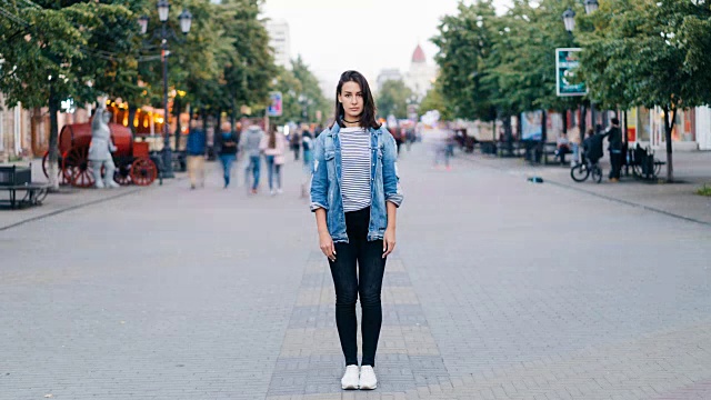 这是一幅压力重重的年轻女子的照片，她穿着牛仔裤和牛仔夹克，独自站在市中心，看着镜头，路人从她身边经过。视频素材