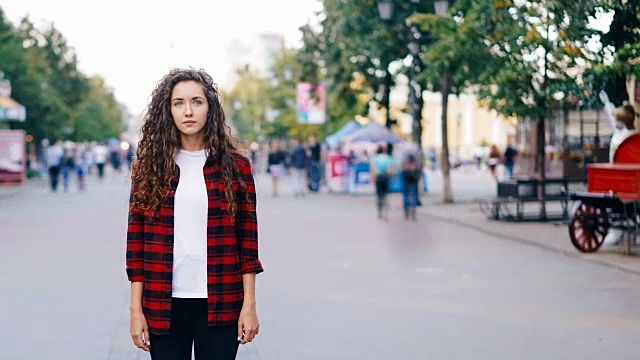 一个孤独的女孩站在市中心的人行道上看着镜头，穿着休闲的衣服，而人群正在四处走动。青春与人生理念。视频下载