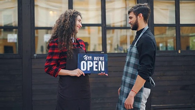 两个快乐的企业家咖啡馆老板的慢动作肖像与开放标志在新建筑前转向相机和微笑欢迎顾客。视频素材