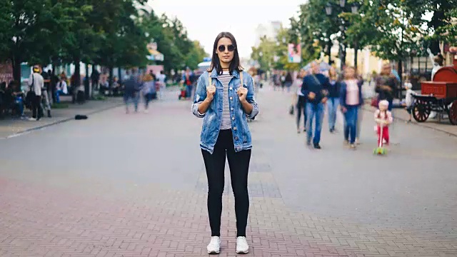 一个迷人的年轻女子戴着太阳镜，背着背包独自站在大城市里，看着镜头，当人群经过。视频素材