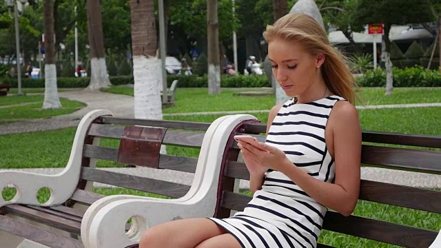 年轻漂亮的苗条女子，一头金色长发，穿着黑白相间的裙子，坐在长椅上，用智能手机拍摄公园的背景。女孩在正方形触摸屏幕上。视频素材