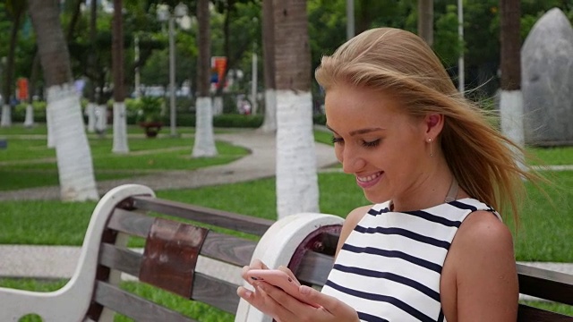 年轻漂亮的苗条女子，一头金色长发，穿着黑白相间的裙子，坐在长椅上，用智能手机拍摄公园的背景。女孩在正方形触摸屏幕上。视频素材