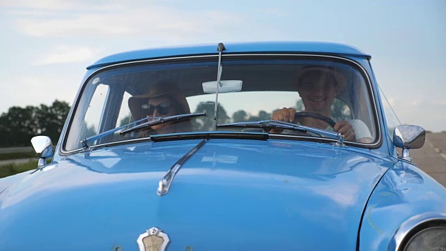 年轻的夫妇在夏天旅行时戴着帽子坐在老式汽车上。快乐的男人和女人坐在老爷车的前座享受旅程和假期。公路旅行的概念。慢动作视频素材