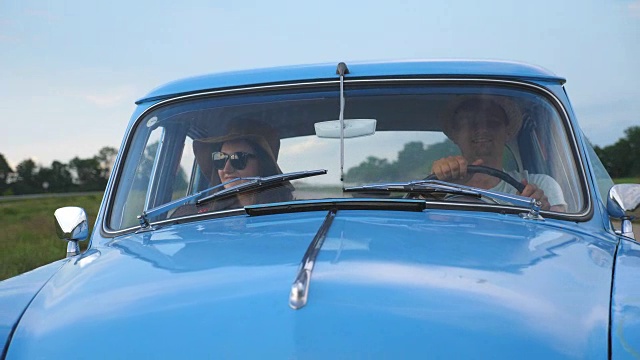 年轻夫妇戴着帽子坐在一辆老爷车里夏天旅行。快乐的男人和女人坐在老爷车的前座享受旅程和假期。公路旅行的概念。慢镜头视频素材