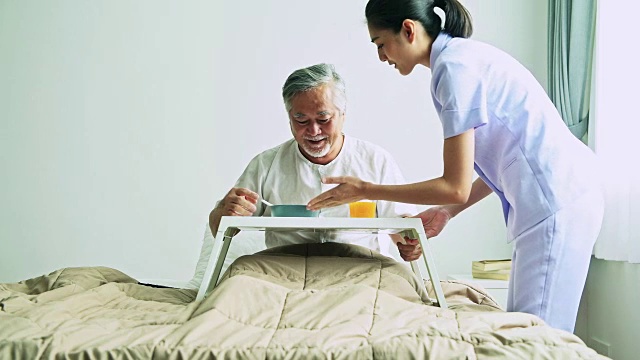 老人躺在床上，一个漂亮的护士正在给他端早餐。年老的亚洲男性与年轻迷人的亚洲女性看管人白胡子。高级家居服务理念。视频下载