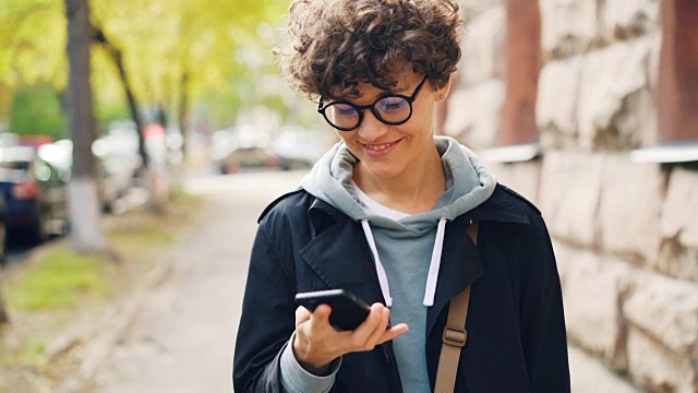 慢镜头微笑的女人戴着眼镜，穿着时髦的衣服，用智能手机看着屏幕，走在城市的户外。青年生活、街头、科技理念。视频素材