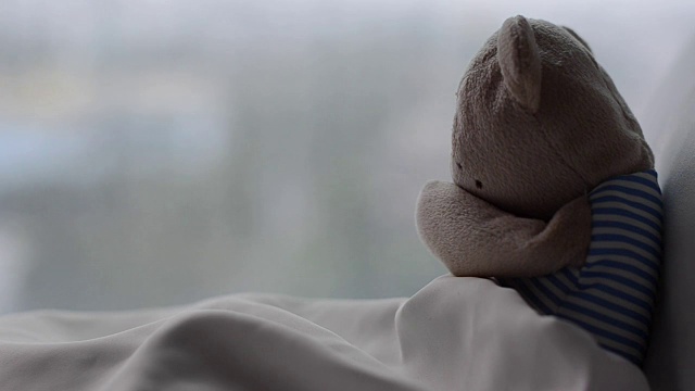 泰迪熊独自在床上哭泣，他的白色枕头和毯子靠近窗户，在早晨的悲伤的一天的雨滴。视频素材