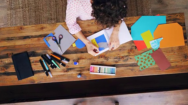 放大的女员工创意设计师在溅纸拼贴在笔记本与蜡笔坐在桌子在现代办公室。工作和创造力的概念。视频素材