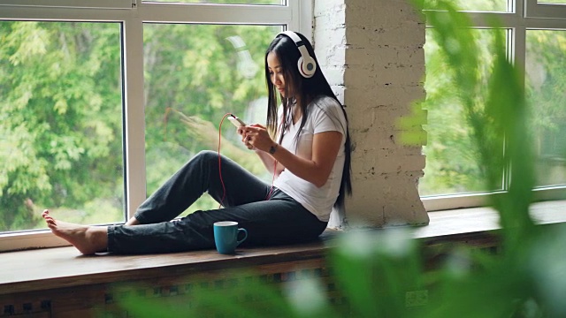 一位快乐的亚洲女性正戴着耳机听着最喜欢的歌曲，坐在窗台上用智能手机微笑和大笑。互联网与青年文化理念。视频素材