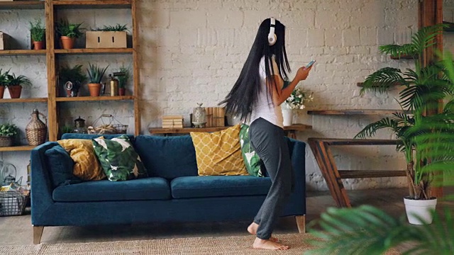 一位迷人的亚洲女士正戴着耳机听音乐，使用智能手机，拿着手机和一杯茶在客厅的地板上跳舞。人与休闲的概念。视频素材
