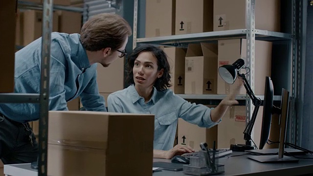 男性和女性仓库库存经理交谈，使用个人电脑和检查库存。在背景的货架上满是纸箱包装。视频素材