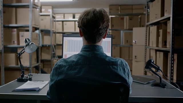 仓库库存经理坐在办公桌前用个人电脑做电子表格。在背景货架上装满了纸箱包装准备运输。视频素材