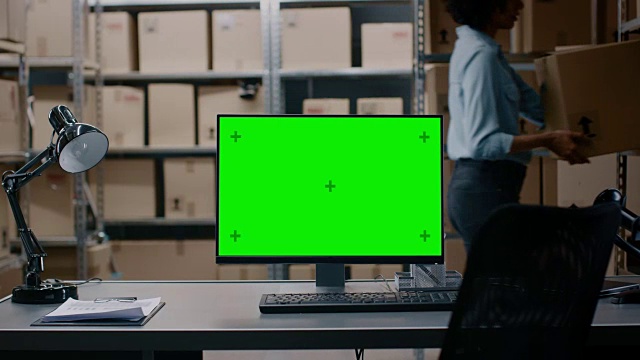 在仓库计算机与绿色模拟屏幕显示站在桌面上。在背景的货架上装满了纸箱和包裹的产品准备运输。视频素材