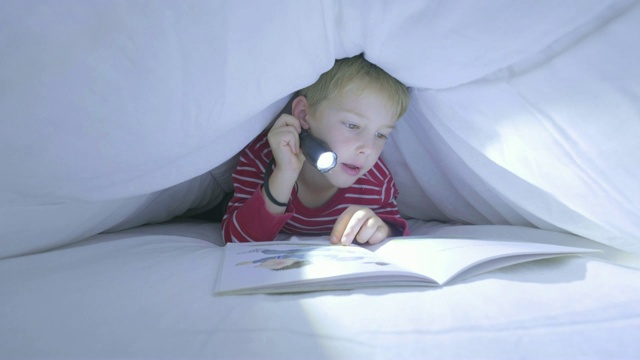 一个白人男孩在羽绒被下用手电筒读故事书视频素材