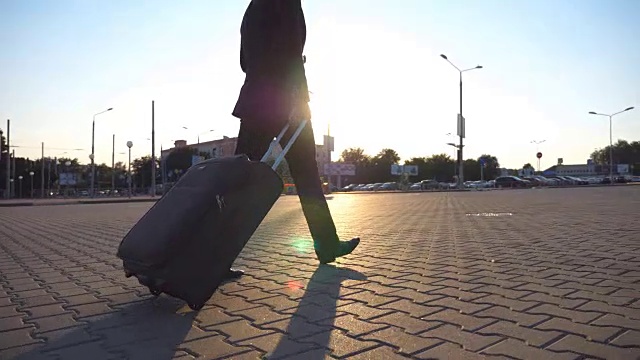 简介成功的商人在一个黑色西装走在他的行李在城市街道日落。一个自信的年轻人拖着带轮子的行李箱走向机场。关闭侧视图视频素材