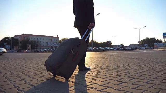 克雷恩拍摄了一个不认识的人走在城市的街道上，在日落时分拉着轮子上的手提箱。年轻的商人看了看手表，拿着行李去机场。后视图视频下载