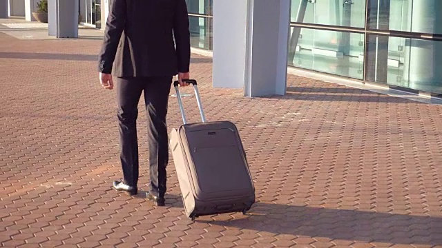 后视图，一个不认识的年轻商人穿着西装走在航站楼附近，拉着轮子上的手提箱。成功的男性商人带着行李去机场。特写慢动作视频素材