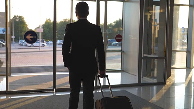 一个面目全非的男人带着他的行李穿过现代机场的玻璃自动门去城市街道。商人从航站楼走到停车场，拖着带轮子的行李箱。后视图慢动作视频下载