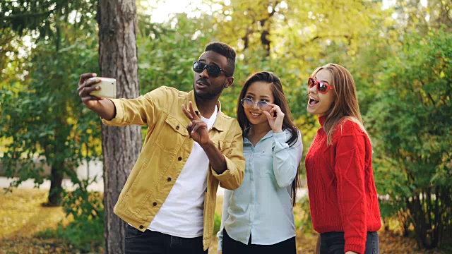 时髦的年轻人在公园里散步，他们戴着太阳镜，微笑着拿着智能手机自拍。人与技术的理念。视频素材