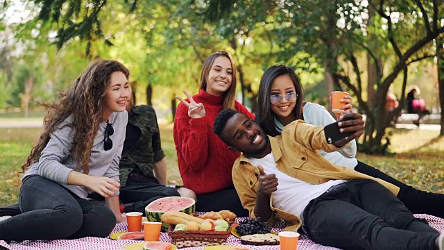 一名快乐的非裔美国人用智能手机与朋友们在野餐时自拍的慢镜头，人们摆着喝饮料的姿势，还做着手势。视频素材