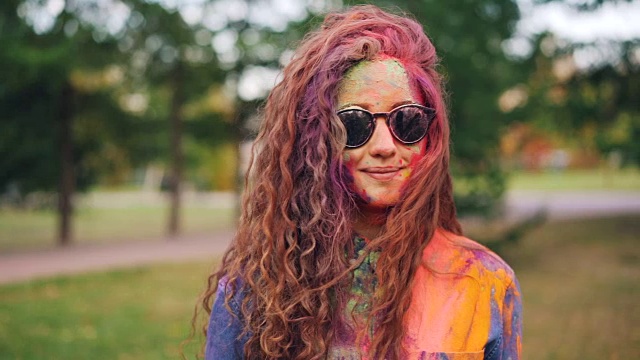 在胡里节，美丽的女孩戴着太阳镜站在户外，脸上和头发上覆盖着明亮的色彩斑斓的红色颜料。青年与传统观念。视频下载