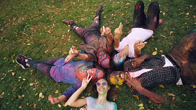 慢动作肖像，放松的人躺在草地上的公园，彩色的脸和衣服，看着相机和微笑移动的手。党与自然观念。视频下载