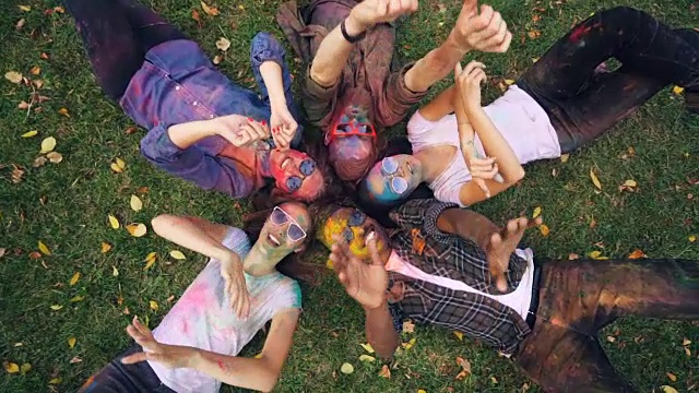 放大轻松的人多民族的群体躺在草地上，一起彩色的脸和衣服，看着相机，微笑和移动的手。放松和聚会的概念。视频下载