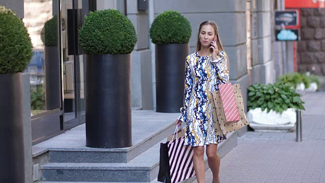 一个穿着长裙的年轻女孩走在城市里购物和讲电话。4 k慢动作视频素材