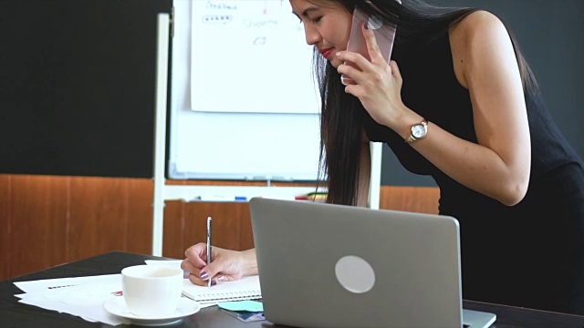 在现代办公室、生活方式和商业理念中，亚洲女商人使用智能手机、用笔记本电脑和咖啡杯在笔记本上写字的4k视频场景视频素材