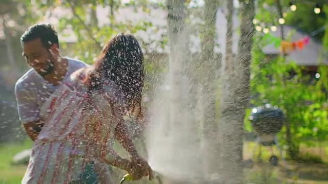 快乐的年轻夫妇在一个炎热的夏天玩水软管洒水器在花园。两个年轻人在爱中湿了开玩笑与软管打架。在缓慢的运动。视频素材