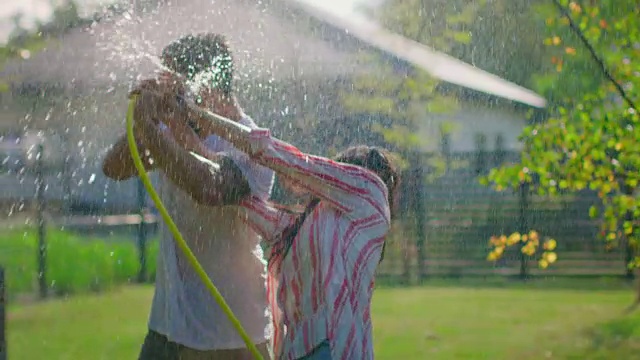 快乐的年轻夫妇在一个炎热的夏天玩水软管洒水器在花园。到处喷水，弄湿了他们。在缓慢的运动。视频素材