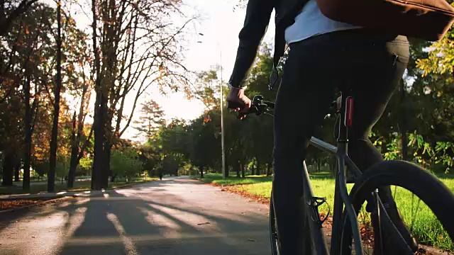 年轻的商人背着皮包骑着自行车去城市公园，慢镜头，后视镜视频下载