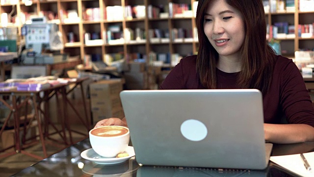 4k视频场景的亚洲女商人工作与笔记本电脑和喝咖啡拿铁艺术杯在咖啡咖啡馆，商业和生活方式的概念视频素材