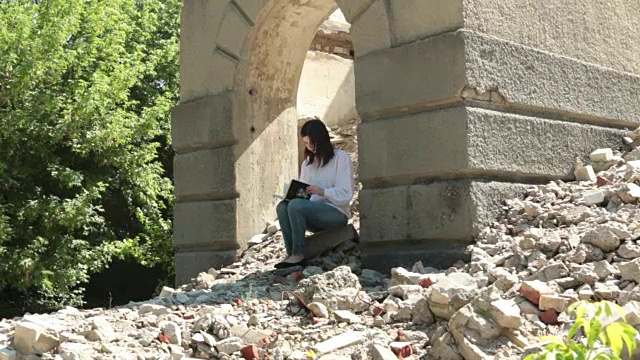 美丽的黑发姑娘坐在一栋建筑物的废墟上看书。视频下载