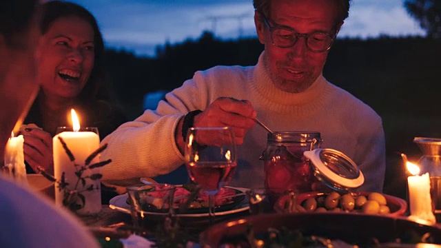 在后院的收获晚宴上，一个兴高采烈的男人一边从餐桌上的罐子里取出腌洋葱圈，一边和朋友聊天视频素材