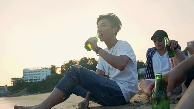 年轻的亚洲成年人悠闲地坐在海滩上喝啤酒视频下载