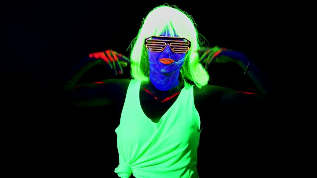 这个女人脸上涂着紫外线，戴着假发，戴着发光眼镜，穿着发光的衣服在镜头前跳舞。白人女人。。视频下载