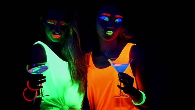 女人脸上涂着紫外线，激光，发光的手镯，饮料，发光的衣服在镜头前一起跳舞，拍摄半身像。白种人和亚洲女人…视频下载