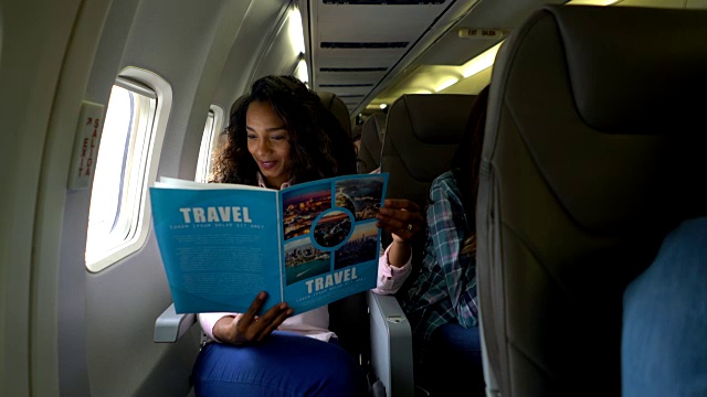 美丽的黑人妇女看着一个旅行指南在飞机上微笑视频下载