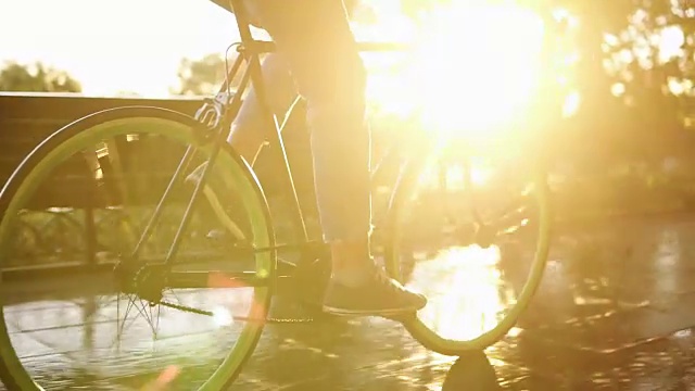 女性的脚在早上的公园或大道骑自行车的近镜头。侧面的一个年轻女子骑着一辆徒步自行车，穿着运动鞋和牛仔裤。阳光照在背景上视频素材