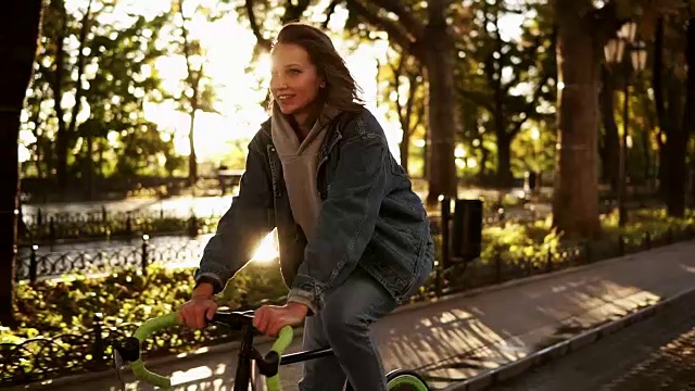 美丽的女人骑自行车在城市铺设的道路。骑自行车的女人早上在公园里骑车。背景上的镜头光晕视频素材