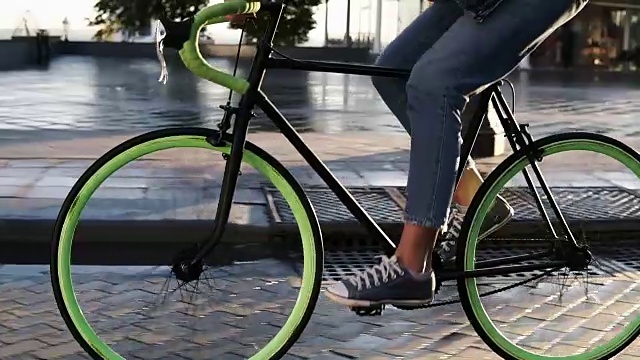 近距离镜头，女性的脚在早上骑自行车的城市街道与潮湿的沥青。一个年轻女子骑着绿色车轮的徒步自行车，穿着运动鞋和牛仔裤的侧视图视频素材