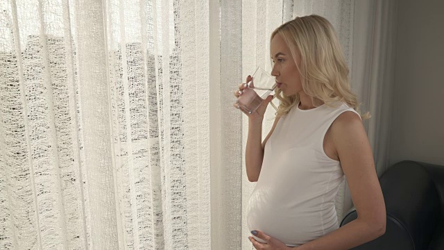 一个怀孕的金发女郎站在被薄纱覆盖的窗户旁的侧视图。她抚摸着自己的肚子，想到什么就笑。她正在喝水，若有所思地看着前面视频素材