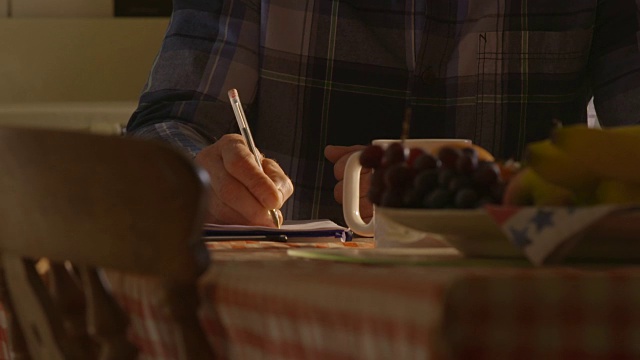 一位中年男子坐在餐桌旁，用红色圆珠笔在信纸上写字。视频素材